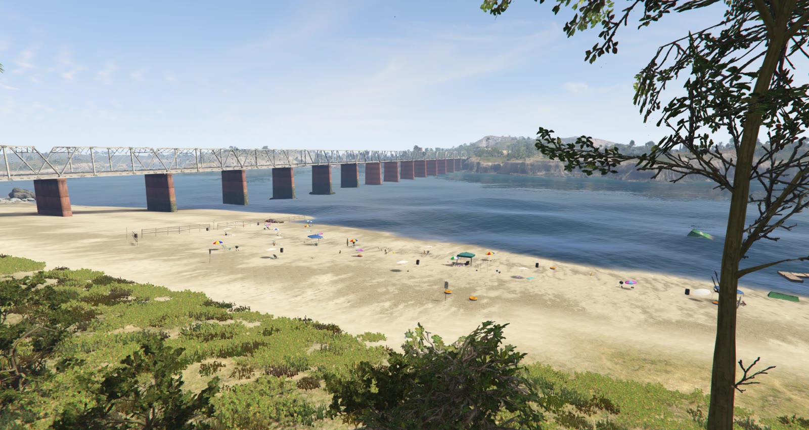 A wide sandy beach beneath a long rail bridge, dotted with colourful parasols, with calm water and green hills beyond