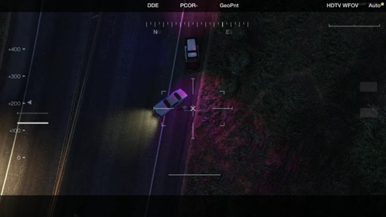 Thermal helicopter camera view looking down on a road at night, with a patrol car and a second car stopped at an angle across the lane